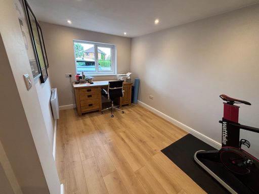A spacious, well-lit room with a desk, chair, and exercise bike against a light wall.